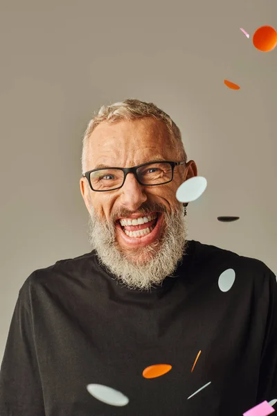 Joyeux modèle masculin mature avec des lunettes souriant joyeusement à la caméra et posant sous la pluie confettis — Photo de stock
