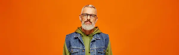 Funky mature man in vibrant hoodie and stylish vest looking at camera on orange backdrop, banner — Stock Photo