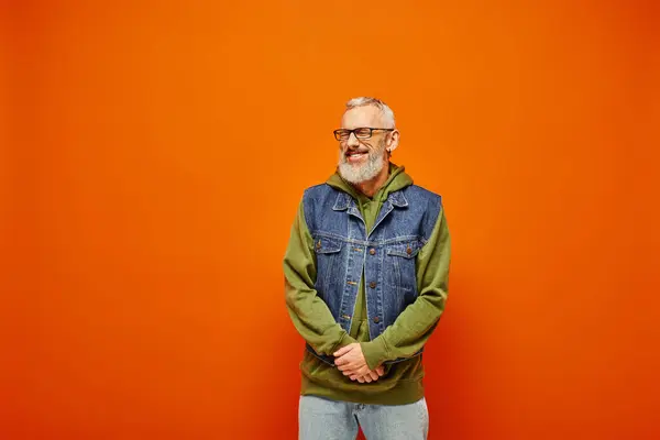 Fröhlich gut aussehender, reifer Mann in grünem, lebendigem Kapuzenpulli mit Jeansweste posiert auf orangefarbenem Hintergrund — Stockfoto