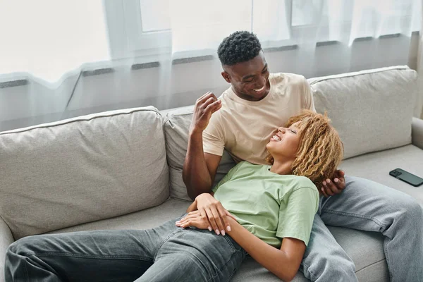 Feliz pareja afroamericana riendo juntos mientras se relaja en el sofá en la sala de estar, alegre - foto de stock