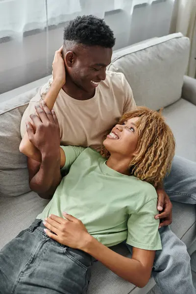 Feliz casal afro-americano rindo enquanto relaxa no sofá, mulher negra deitada em suas voltas — Fotografia de Stock