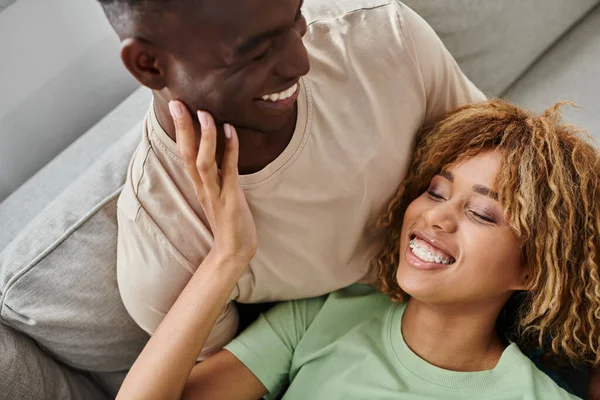 African american couple laughing while resting on sofa, cheerful woman lying on laps of boyfriend — Stock Photo