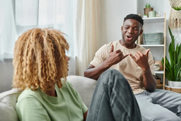 Alegre hombre negro sonriendo mientras se comunica con el lenguaje de señas con la novia en la sala de estar - foto de stock
