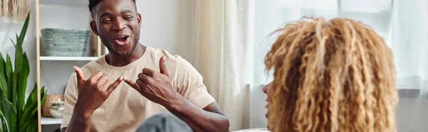 Радісний чорний чоловік посміхається під час спілкування з жестовою мовою з подругою вдома, банер — стокове фото