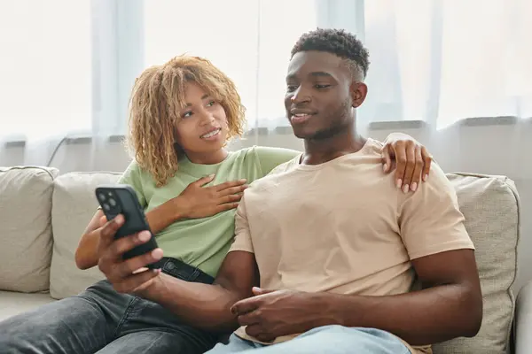 Счастливая африканская американка в брекетах обнимается и смотрит на мужчину со смартфоном, общение — стоковое фото