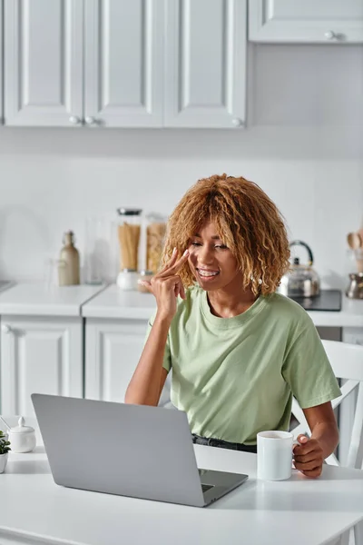 Heureuse femme afro-américaine en utilisant le langage des signes lors d'un appel vidéo et tenant une tasse de café — Photo de stock