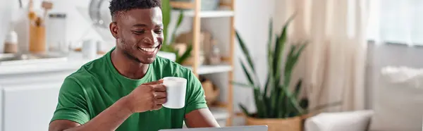 Sonriente hombre afroamericano disfrutando de una taza de café y sonriendo en la cocina, pancarta horizontal - foto de stock