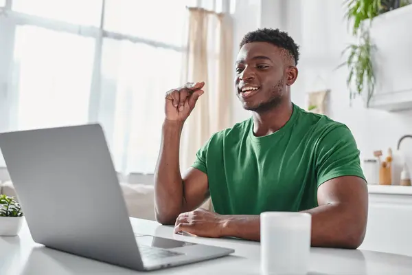 Hombre afroamericano enfocado sonriendo mientras se utiliza el lenguaje de señas durante la videollamada en el portátil en casa - foto de stock
