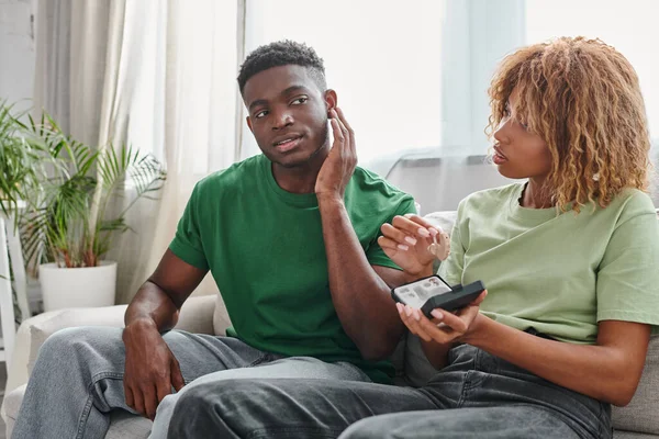 African american man touching ear near girlfriend holding medical device in hands, hearing aid — Stock Photo