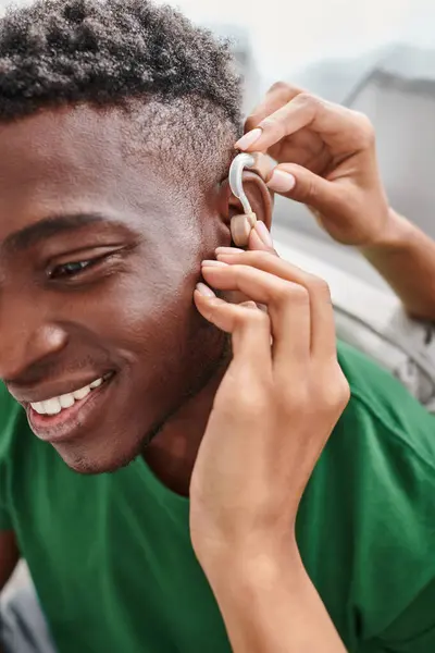 Счастливый африканский американец улыбается, когда его подруга помогает со слуховым аппаратом, медицинским оборудованием — стоковое фото