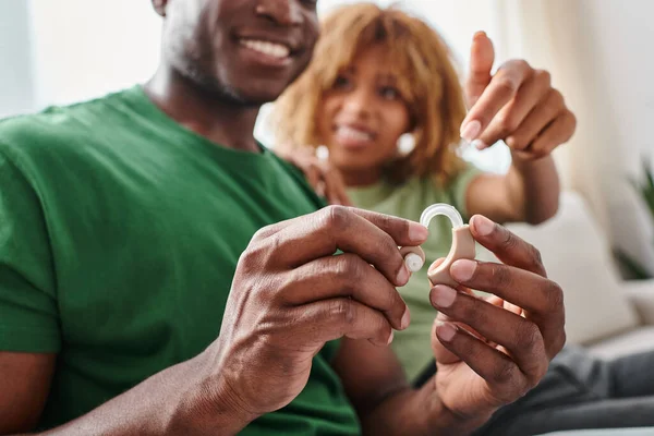 Fokus auf Hörgerät, glücklicher afrikanisch-amerikanischer Mann hält Medizinprodukt in der Nähe seiner Freundin — Stockfoto