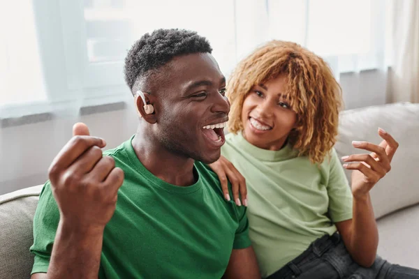 Hombre afroamericano emocionado en dispositivo de audífono sentado cerca de novia feliz en aparatos ortopédicos - foto de stock