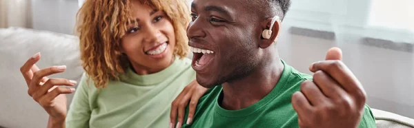 Excited african american man in hearing aid device sitting near happy girlfriend in braces, banner — Stock Photo