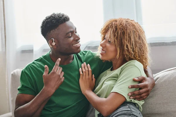 Joyeux couple afro-américain embrassant sur le canapé, sourd homme noir dans l'appareil auditif et femme dans les bretelles — Photo de stock