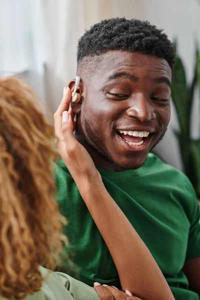 African american woman wearing hearing aid medical device on ear of excited boyfriend, accessibility — Stock Photo