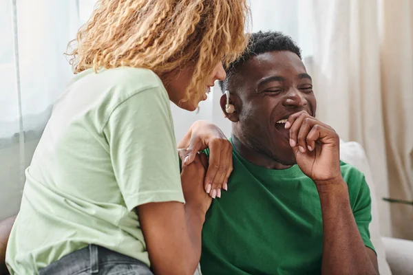 Hombre afroamericano excitado en dispositivo de audífono sonriendo felizmente cerca de su novia en casa - foto de stock
