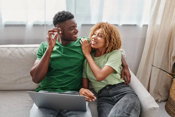 Felice uomo africano americano in apparecchio acustico seduto sul divano con la fidanzata e utilizzando il computer portatile — Foto stock
