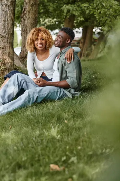 Heureux couple afro-américain en tenue décontractée étreinte tout en étant assis ensemble sur l'herbe, se tenant la main — Photo de stock
