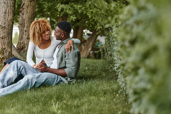 Heureuse femme afro-américaine en tenue décontractée étreignant petit ami tout en étant assis ensemble sur l'herbe — Photo de stock