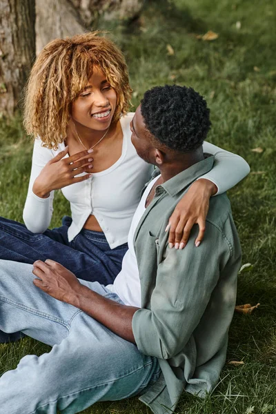 Mujer afroamericana feliz en ropa casual mirando al novio y sentados juntos en la hierba - foto de stock