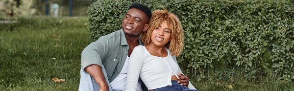 Cheerful african american couple hugging and sitting together on green grass, happiness banner — Stock Photo
