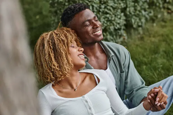 Excitée femme afro-américaine dans les bretelles tenant la main avec petit ami tout en étant assis dans le parc — Photo de stock