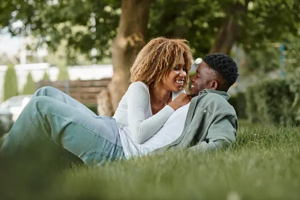 Joyeux jeune couple afro-américain partageant un regard affectueux tout en étant assis sur une herbe dans le parc — Photo de stock