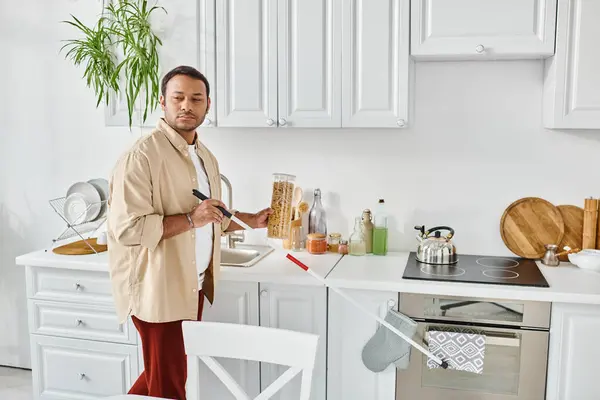 Atractivo hombre indio con ceguera en traje casual recoger tarro de macarrones mientras está en la cocina - foto de stock