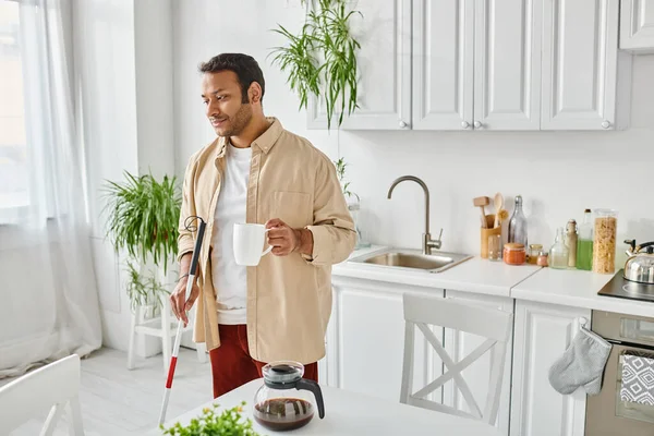 Красивий індійський чоловік з обмеженими можливостями в затишному домашньому одязі з палицею, що тримає чашку кави на кухні — стокове фото