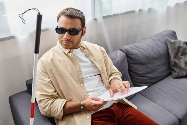 Good looking indian man with blindness in glasses sitting on sofa and reading braille code — Stock Photo