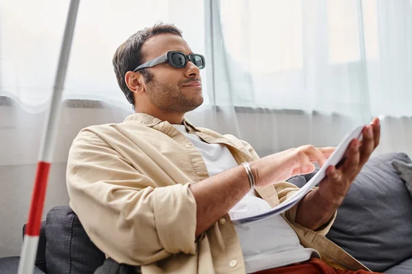 Fröhlicher indischer Mann mit Sehbehinderung mit Brille auf Sofa sitzend und Brailleschrift lesend — Stockfoto