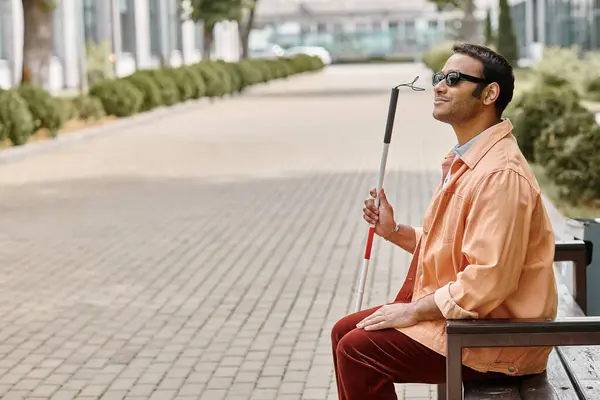Glad indian blind man in orange jacket sitting outside on bench with walking stick and glasses — Stock Photo