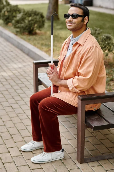 Веселый индийский слепой в оранжевой куртке сидит снаружи на скамейке с тростью и очками — стоковое фото