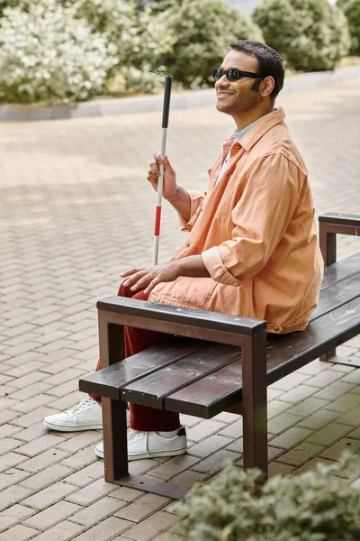 Радісний індійський сліпий чоловік в помаранчевій куртці сидить на лавці з палицею і окулярами — стокове фото