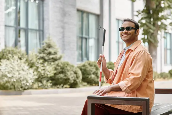 Fröhlicher indischer Blinder in orangefarbener Jacke sitzt draußen auf Bank mit Gehstock und Brille — Stockfoto