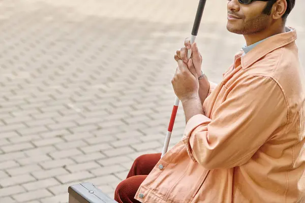 Cheerful indian blind man in orange jacket sitting outside on bench with walking stick and glasses — Stock Photo