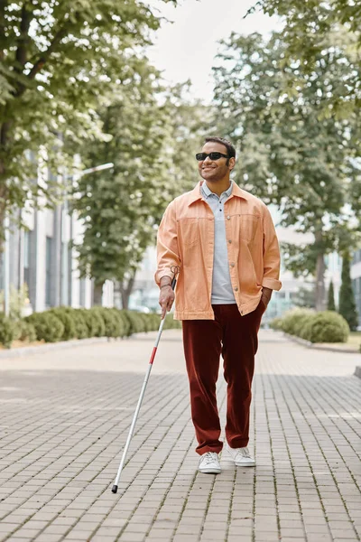 Joyful indian blind man with glasses and walking stick in orange vivid jacket walking in park — Stock Photo