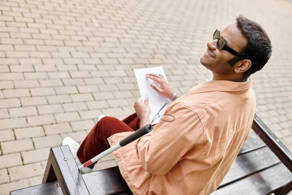 Cheerful indian blind man in orange jacket sitting on bench and reading braille code while outside — Stock Photo