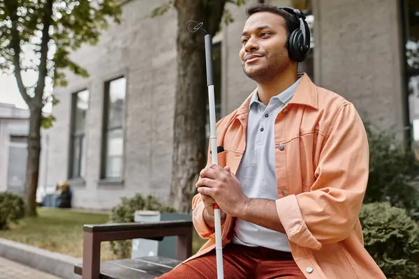 Cheerful indian man with blindness in headphones with walking stick enjoying music while outside — Stock Photo