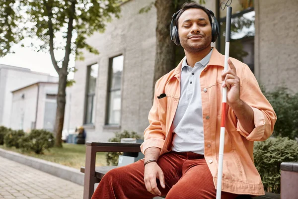 Joyous indian man with blindness in headphones with walking stick enjoying music while outside — Stock Photo