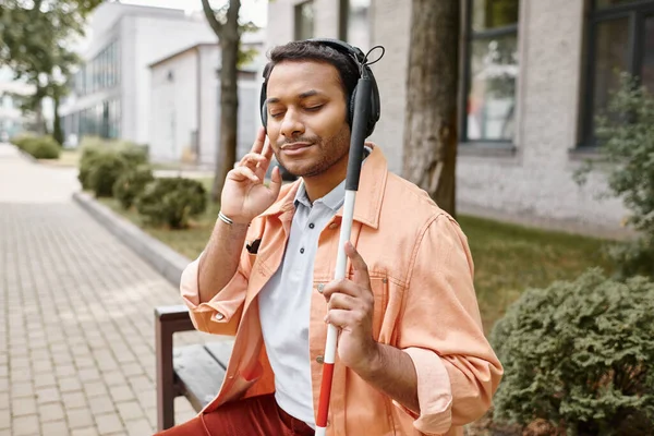 Merry indian man with blindness in headphones with walking stick enjoying music while outside — Stock Photo