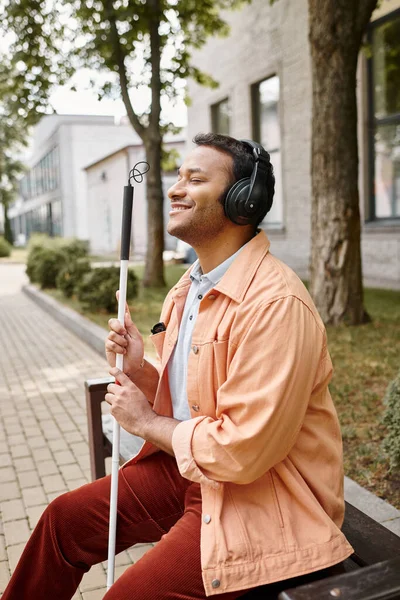 Alegre homem indiano com deficiência em roupa casual com fones de ouvido e bengala desfrutando de música — Fotografia de Stock