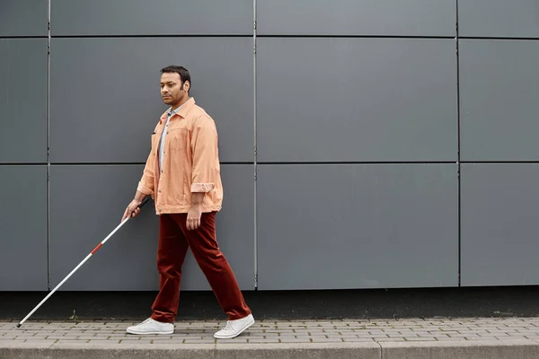 Schöner indisch blinder Mann in orangefarbener Jacke mit helfendem Stock beim Gehen mit grauer Wand vor Hintergrund — Stockfoto