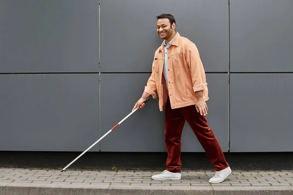 Cheerful indian blind man in orange jacket with helping stick walking with gray wall on backdrop — Stock Photo