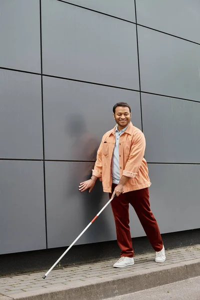 Fröhlicher indisch blinder Mann in orangefarbener Jacke mit helfendem Stock beim Gehen mit grauer Wand vor Hintergrund — Stockfoto
