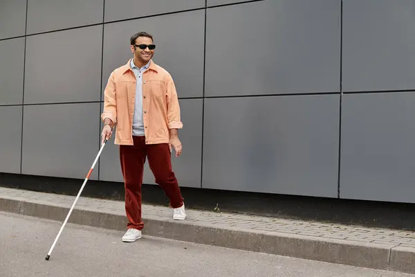 Fröhlicher indischer Blinder in orangefarbener Jacke mit Spazierstock und Brille mit grauer Wand auf Hintergrund — Stockfoto