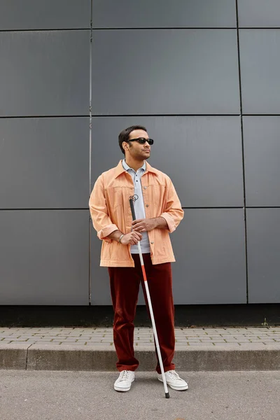 Gut aussehender indischer Behinderter in orangefarbener Jacke mit Brille und Stock mit grauer Wand auf Hintergrund — Stockfoto