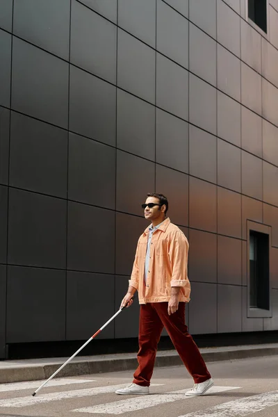 Attraktiver indischer Blinder in orangefarbener Jacke mit Brille und Stock mit grauer Wand auf Hintergrund — Stockfoto