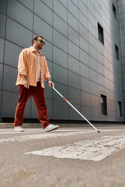 Optimistischer indischer Blinder in orangefarbener Jacke mit Spazierstock und Brille mit grauer Wand auf Hintergrund — Stockfoto