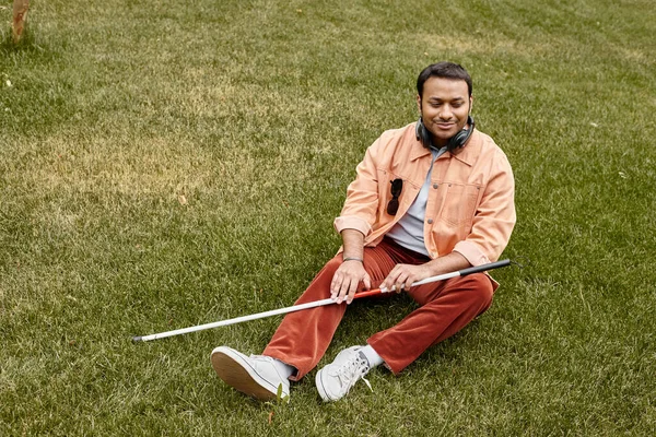 Cheerful indian blind man in orange jacket sitting on grass with headphones and walking stick — Stock Photo
